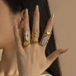 Tanned hand wearing LIVKE shell ring layered with thin gold bands against ocean waves