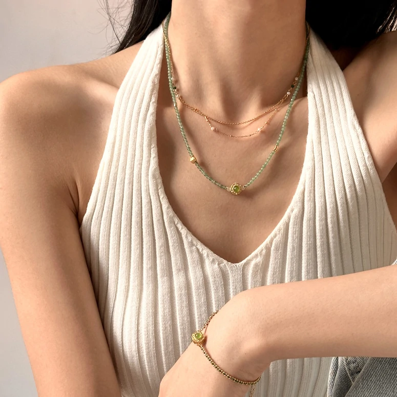 Adjustable layered bracelet with green braided cord, gold bead chains, and olive stone pendant on linen fabric, showing hypoallergenic clasp and boho styling for daily wear