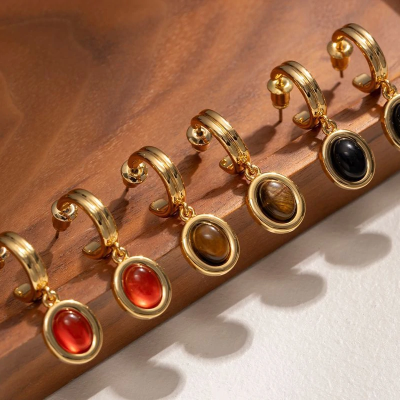 Layered 18K gold-plated tiger eye earrings with red/black/brown stone pendants on terrazzo backdrop, styled with leather cord necklaces